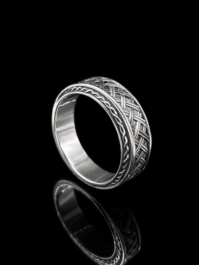 A cinematic studio shot of a wide sterling silver ring lying on a reflective golden surface. The band features a central "basket weave" pattern—diagonal strands of silver crossing over and under each other. The edges are framed by a distinct "rope" or "cable" twist border. The raised metal is polished silver, while the deep recesses are blackened to highlight the intricate weaving.