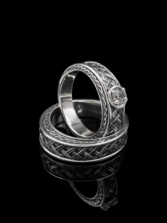 A cinematic studio shot of a wide sterling silver ring lying on a reflective golden surface. The band features a central "basket weave" pattern—diagonal strands of silver crossing over and under each other. The edges are framed by a distinct "rope" or "cable" twist border. The raised metal is polished silver, while the deep recesses are blackened to highlight the intricate weaving.