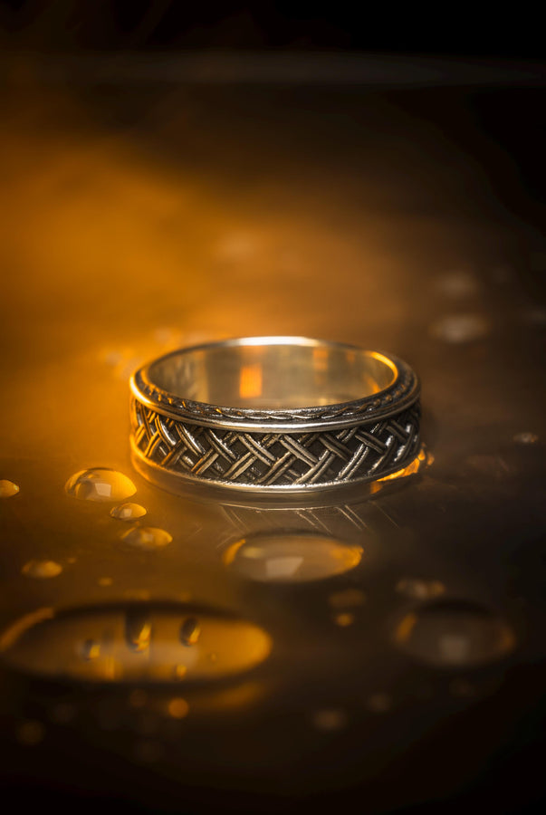 A cinematic studio shot of a wide sterling silver ring lying on a reflective golden surface. The band features a central "basket weave" pattern—diagonal strands of silver crossing over and under each other. The edges are framed by a distinct "rope" or "cable" twist border. The raised metal is polished silver, while the deep recesses are blackened to highlight the intricate weaving.