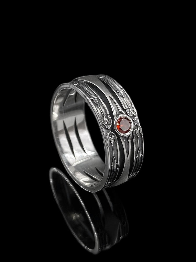 A detailed studio shot of a wide, heavy-gauge sterling silver ring. The focal point is a round, deep red gemstone (Garnet) bezel-set in the center. Flanking the stone are three parallel, tapered grooves that resemble the fletching of an arrow or the futuristic paneling of armor. The metal surfaces are brushed silver, while the deep grooves are blackened with oxidation to emphasize the directional lines. (etsy image id: 7441158070)