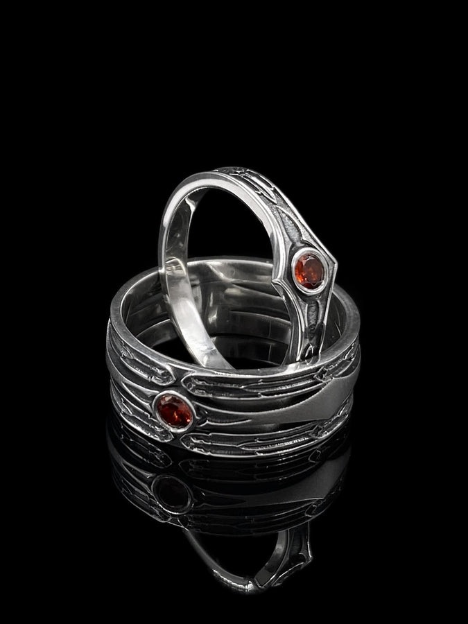A detailed studio shot of a wide, heavy-gauge sterling silver ring. The focal point is a round, deep red gemstone (Garnet) bezel-set in the center. Flanking the stone are three parallel, tapered grooves that resemble the fletching of an arrow or the futuristic paneling of armor. The metal surfaces are brushed silver, while the deep grooves are blackened with oxidation to emphasize the directional lines. (etsy image id: 7441158070)