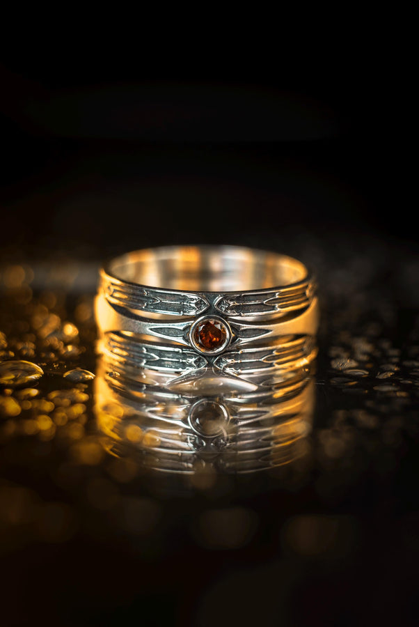 A detailed studio shot of a wide, heavy-gauge sterling silver ring. The focal point is a round, deep red gemstone (Garnet) bezel-set in the center. Flanking the stone are three parallel, tapered grooves that resemble the fletching of an arrow or the futuristic paneling of armor. The metal surfaces are brushed silver, while the deep grooves are blackened with oxidation to emphasize the directional lines. (etsy image id: 7441158070)