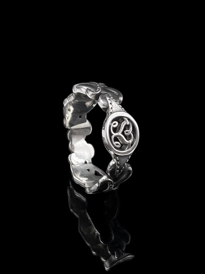 A cinematic macro shot of a sterling silver ring bathed in warm, golden light. The focal point is the central oval face, which encloses a raised relief design of two spirals meeting in the center to suggest a heart shape. The band shoulders feature textured knotwork that leads up to the centerpiece. The ring rests on a wet, reflective surface, creating a mirror image of the love knot below. (etsy image id: 7490345549)