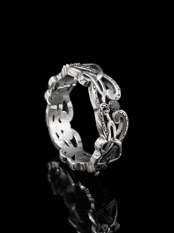 A cinematic close-up of a sterling silver ring lying on a reflective surface. The band features an "openwork" design, meaning the Celtic knot pattern is cut out, allowing light to pass through the loops. The silver strands weave over and under in a continuous infinity motif. The background is dark, but the ring is illuminated by a warm, golden mist. (etsy image id: 7442429936)