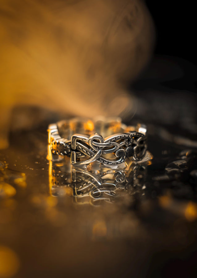 A cinematic close-up of a sterling silver ring lying on a reflective surface. The band features an "openwork" design, meaning the Celtic knot pattern is cut out, allowing light to pass through the loops. The silver strands weave over and under in a continuous infinity motif. The background is dark, but the ring is illuminated by a warm, golden mist. (etsy image id: 7442429936)