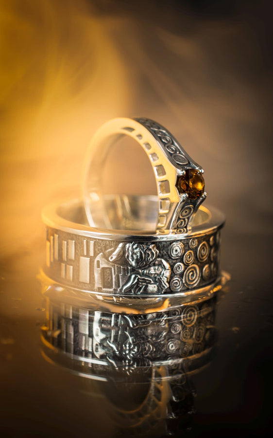 A detailed macro shot of a sterling silver engagement ring. The focal point is a round, honey-colored gemstone (Citrine or Madeira Topaz) set flush into the band. The shank is decorated with a continuous relief pattern of spirals and circles, mimicking the decorative robes found in Gustav Klimt's "Golden Phase" paintings. The silver features an oxidized finish, creating a dark background that makes the polished spirals pop. (etsy image id: 7142785932)