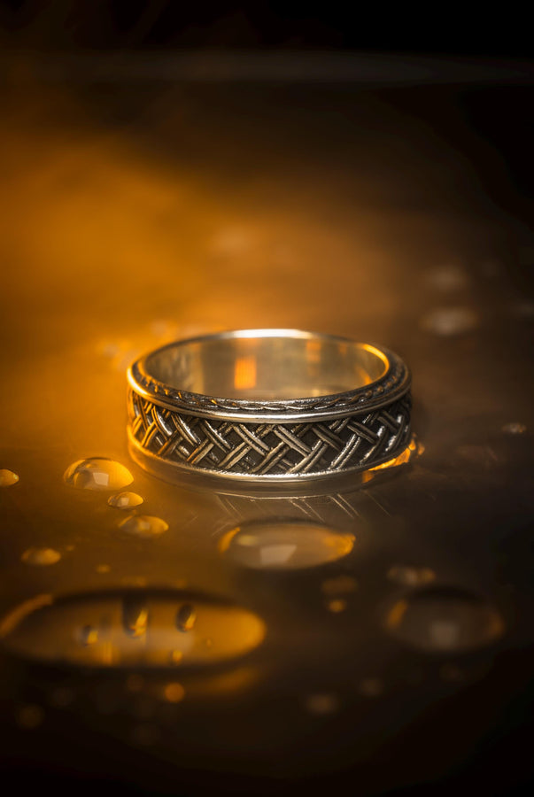 A warm, atmospheric shot of a matching wedding ring set. The engagement ring stands upright, showcasing a round white gemstone in a bezel setting. Lying flat beneath it is a matching wide band without a stone. Both rings share a cohesive design: a central diagonal "basket weave" pattern flanked by twisted rope borders. The silver is oxidized to reveal the intricate texture of the weave.