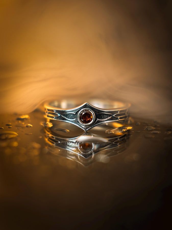 A cinematic close-up of a sterling silver ring with a distinct "V" shaped or "Lance Tip" contour. The focal point is a bezel-set, round gemstone with a deep amber-orange hue (Citrine or Madeira Citrine). The shoulders of the band feature horizontal grooves that taper toward the center, resembling the shaft of a spear or the fletching of an arrow. The metal is polished but features dark oxidation in the grooves for contrast. (etsy image id: 7489049399)