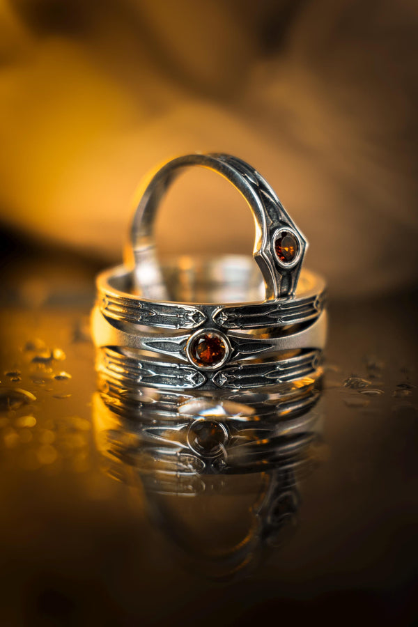 A cinematic close-up of a sterling silver ring with a distinct "V" shaped or "Lance Tip" contour. The focal point is a bezel-set, round gemstone with a deep amber-orange hue (Citrine or Madeira Citrine). The shoulders of the band feature horizontal grooves that taper toward the center, resembling the shaft of a spear or the fletching of an arrow. The metal is polished but features dark oxidation in the grooves for contrast. (etsy image id: 7489049399)