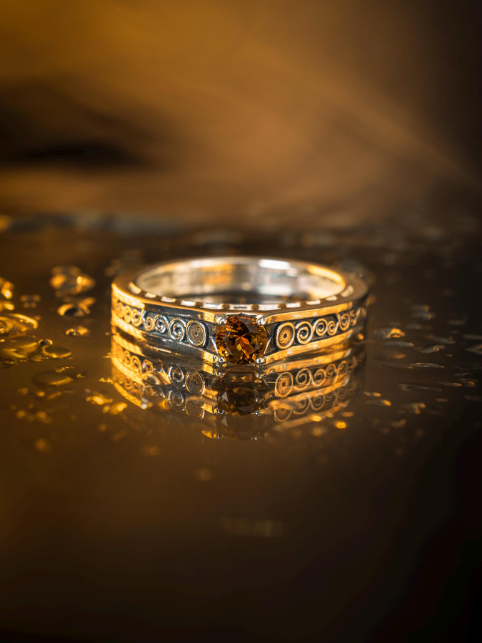 A cinematic studio shot of a matching two-piece ring set. The bottom ring is a wide silver band featuring a detailed relief carving of the couple from Gustav Klimt's "The Kiss." The top ring is a narrower engagement band that mimics the geometric patterns of the painting and holds a round, amber-colored gemstone. The background is a warm, golden haze, reflecting the "Golden Phase" of the original artwork.