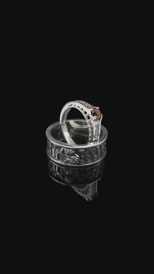 A cinematic studio shot of a matching two-piece ring set. The bottom ring is a wide silver band featuring a detailed relief carving of the couple from Gustav Klimt's "The Kiss." The top ring is a narrower engagement band that mimics the geometric patterns of the painting and holds a round, amber-colored gemstone. The background is a warm, golden haze, reflecting the "Golden Phase" of the original artwork.
