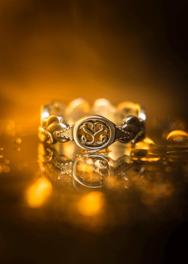 A cinematic macro shot of a sterling silver ring bathed in warm, golden light. The focal point is the central oval face, which encloses a raised relief design of two spirals meeting in the center to suggest a heart shape. The band shoulders feature textured knotwork that leads up to the centerpiece. The ring rests on a wet, reflective surface, creating a mirror image of the love knot below. (etsy image id: 7490345549)