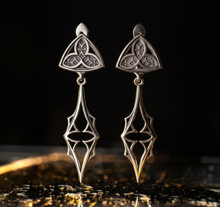 Embrace your dark beauty with our Gothic silver earrings. Unique design, premium silver, intricate details, and comfortable to wear.
A sharp studio shot of a pair of articulated silver earrings. The top stud is a soft triangle shape containing a raised "trefoil" or "trinity" knot pattern against a textured background. The bottom section is an elongated, openwork drop that tapers to a sharp point, resembling a Gothic spire or a (etsy image id: 7218877462)