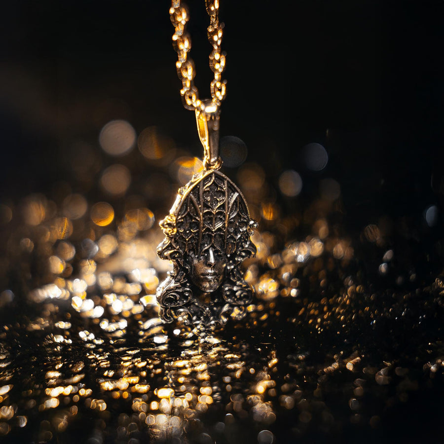 A detailed studio shot of a sterling silver pendant resting on a dark, textured stone surface. The design features a realistic human face in the center, but the forehead and eyes are obscured by a grid of Gothic window tracery (vertical bars and trefoils). The chin is framed by swirling Art Nouveau-style acanthus leaves. The silver is heavily oxidized, giving the face the appearance of a weathered stone statue. (etsy image id: 7448678222)