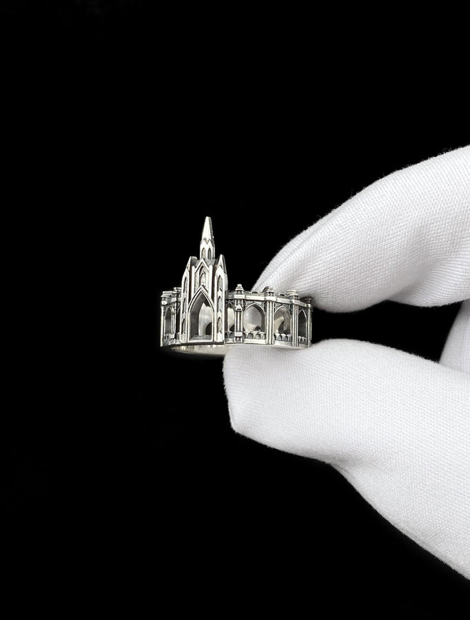 A cinematic studio shot of a sculptural sterling silver ring standing on a reflective surface. The ring is shaped like a miniature building, featuring a tall central spire that rises to a sharp point. The band itself is formed by a colonnade of pointed Gothic arches and buttresses that wrap around the finger. The silver is heavily oxidized, creating deep shadows in the "windows" and "doorways" of the structure. (etsy image id: 7450125356)