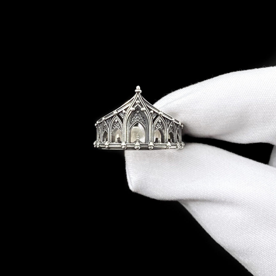 A cinematic studio shot of a sculptural sterling silver ring standing on a reflective surface. The ring is designed as a "crown" or "coronet," formed by a continuous circle of Gothic cathedral arches. The central arch is the tallest, creating a focal point like a spire. The silver is heavily oxidized, with black recesses emphasizing the ribbed vaulting and architectural details against the polished ridges.
