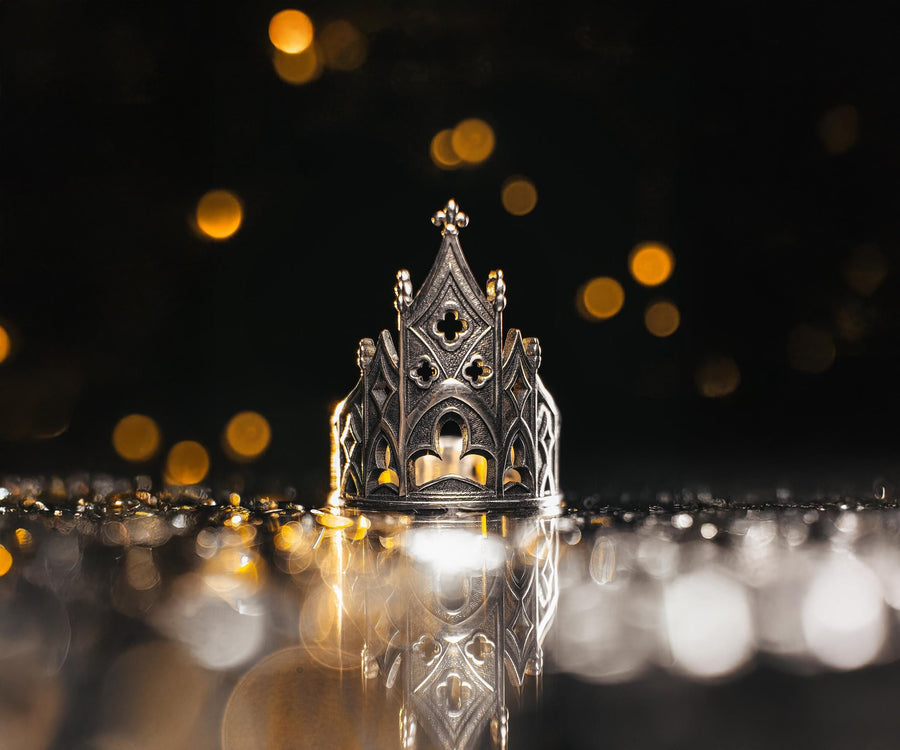 A clean studio shot of a sculptural sterling silver ring resting on a matte grey surface. The ring depicts a Gothic building facade with a tall, triangular central peak (gable) filled with geometric cross-shaped tracery. Flanking the center are lower pointed arches and vertical buttresses that wrap around the band. The silver is matte-finished with dark oxidation in the recesses to mimic stone shadows. (etsy image id: 7452370200)