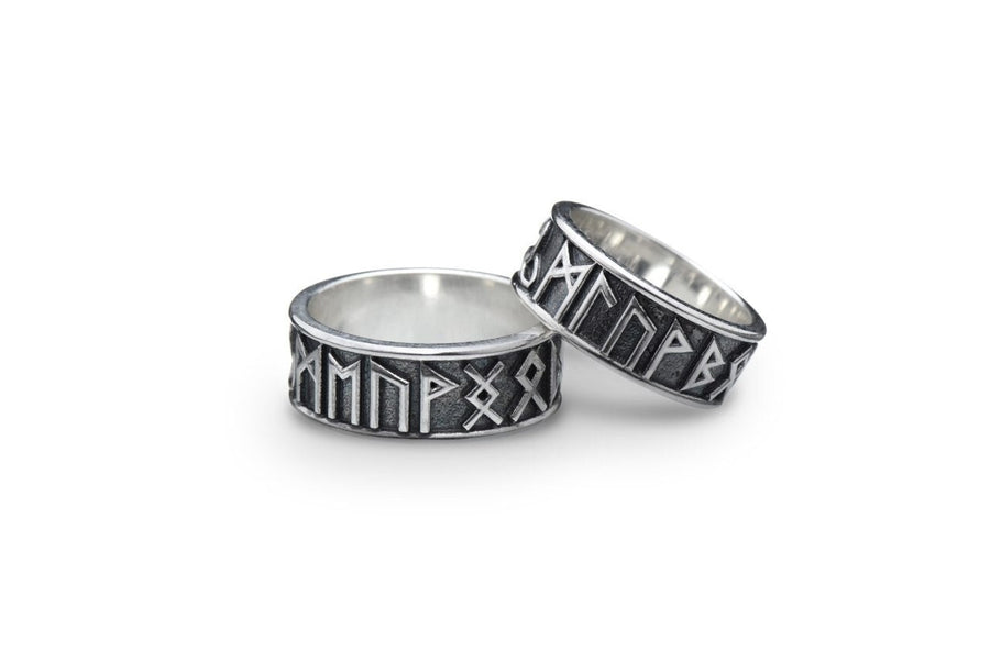 A clean studio shot of two matching sterling silver rings. The bands feature the 24 characters of the Elder Futhark runic alphabet wrapping around the exterior. The runes are polished silver, standing out sharply against a rough, blackened background texture. The rims are smooth and polished, framing the ancient script.