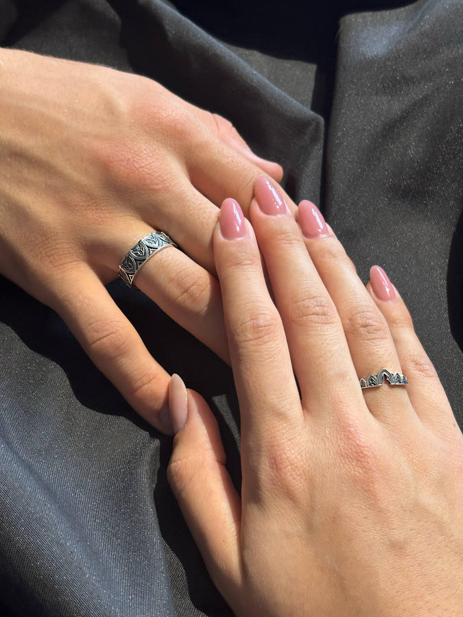 A cinematic studio shot of two matching sterling silver rings. The top ring is a delicate "crown" or tiara shape, formed by the peak of a Gothic arch with trefoil tracery. The bottom ring is a wide band featuring a continuous row of identical arches. The background metal inside the arches has a rough, "eroded" or stippled texture, heavily oxidized to black, while the raised architectural lines are polished smooth.
