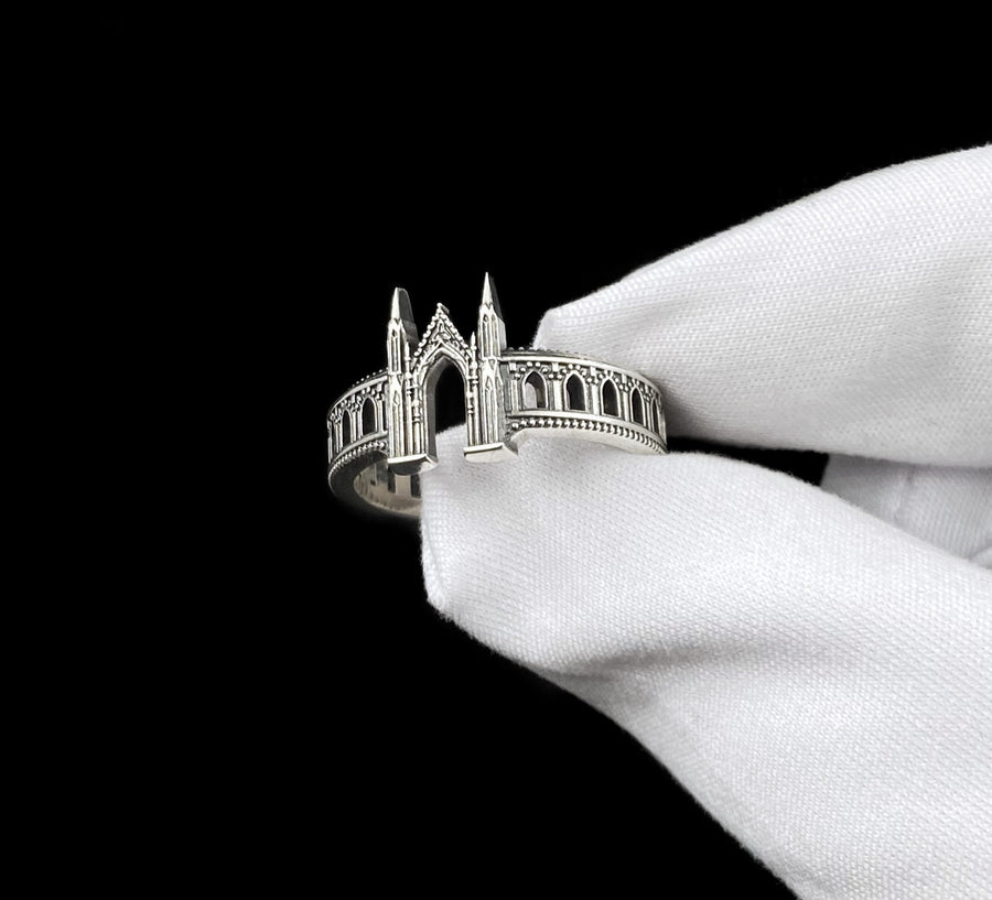A cinematic studio shot of a sculptural sterling silver ring standing on a reflective surface. The design mimics the facade of a Gothic cathedral or castle gate. Two tall, pointed spires rise up on either side of a central archway. The band continues outwards with a colonnade of smaller arches. The silver features a beaded "cornice" detail along the edges and is heavily oxidized to emphasize the depth of the "stonework." (etsy image id: 7450300962)