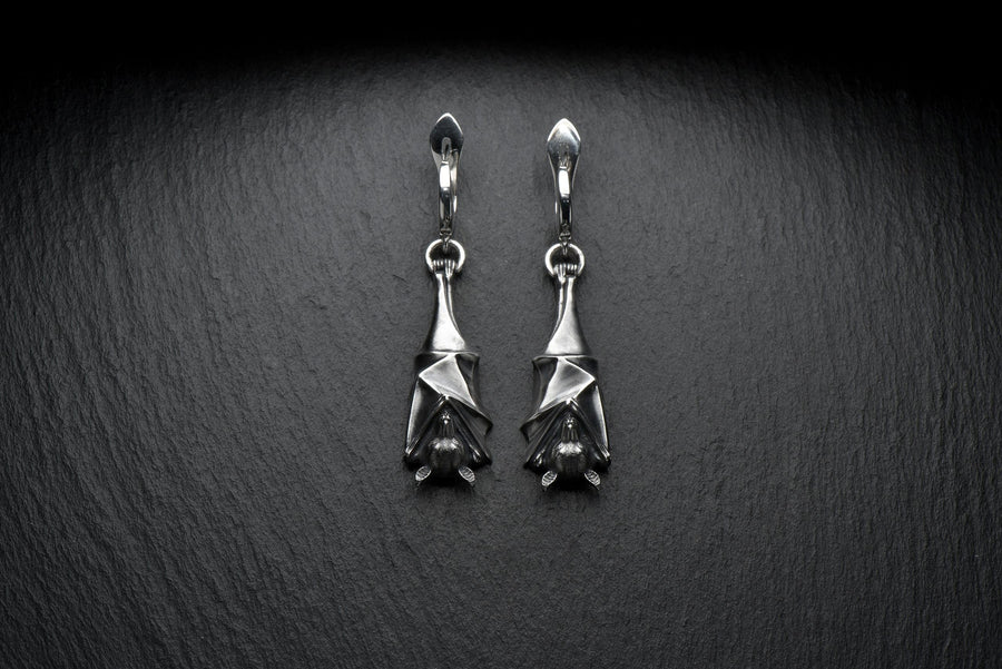 May include: A pair of silver-colored bat-shaped earrings. Each earring features a detailed bat design with outstretched wings and a small loop for attachment. The earrings are suspended from a curved hook. The background is a dark, textured surface. A detailed close-up studio shot of a pair of sterling silver dangle earrings shaped like flying foxes. Each earring features a highly textured sleeping fruit bat hanging upside down, wrapped securely (etsy image id: 7368202736)