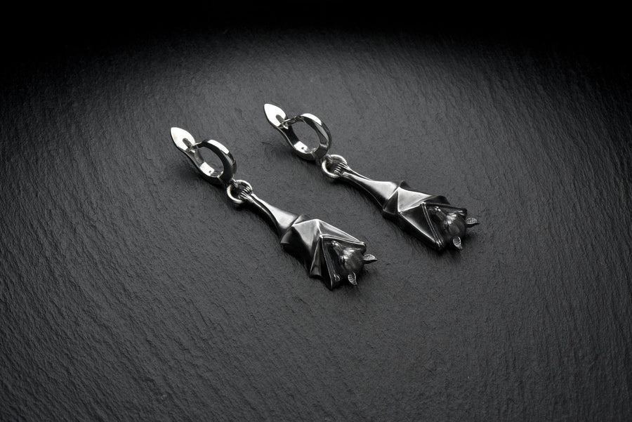 May include: A pair of silver-colored bat-shaped earrings. Each earring features a detailed bat design with outstretched wings and a small loop for attachment. The earrings are suspended from a curved hook. The background is a dark, textured surface. A detailed close-up studio shot of a pair of sterling silver dangle earrings shaped like flying foxes. Each earring features a highly textured sleeping fruit bat hanging upside down, wrapped securely (etsy image id: 7368202736)