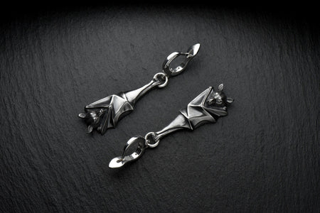 May include: A pair of silver-colored bat-shaped earrings. Each earring features a detailed bat design with outstretched wings and a small loop for attachment. The earrings are suspended from a curved hook. The background is a dark, textured surface. A detailed close-up studio shot of a pair of sterling silver dangle earrings shaped like flying foxes. Each earring features a highly textured sleeping fruit bat hanging upside down, wrapped securely (etsy image id: 7368202736)