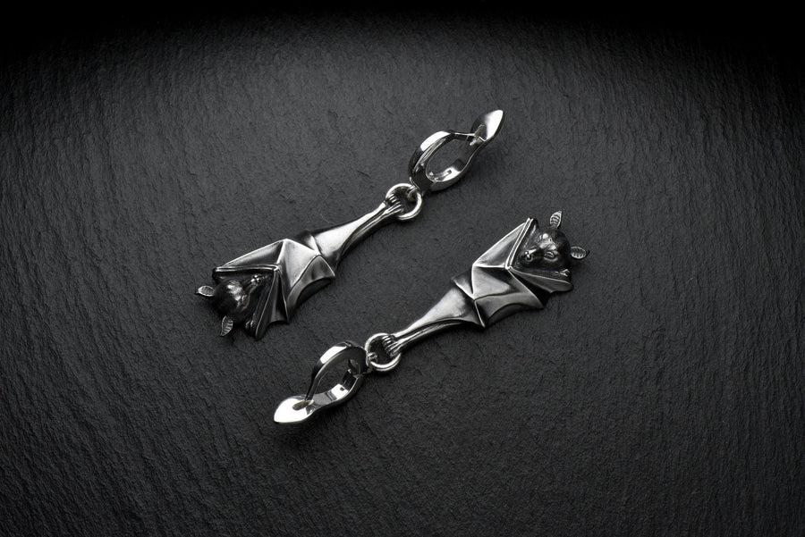 May include: A pair of silver-colored bat-shaped earrings. Each earring features a detailed bat design with outstretched wings and a small loop for attachment. The earrings are suspended from a curved hook. The background is a dark, textured surface. A detailed close-up studio shot of a pair of sterling silver dangle earrings shaped like flying foxes. Each earring features a highly textured sleeping fruit bat hanging upside down, wrapped securely (etsy image id: 7368202736)