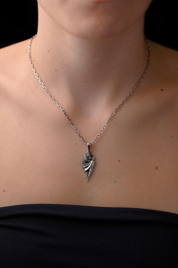 A detailed close-up studio shot of a sterling silver pendant shaped like two flying foxes or fruit bats. The bats are intimately huddled together, wrapped within the smooth, sweeping folds of their leathery wings. The metal is highly textured to show the fine details of their fur and faces. The silver is heavily oxidized in the recesses, creating a striking contrast against the polished highlights of the wings. (etsy image id: 5592925539)