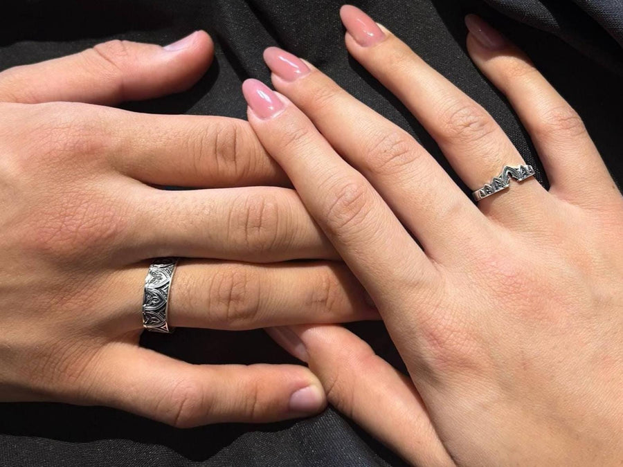A cinematic studio shot of two matching sterling silver rings. The top ring is a delicate "crown" or tiara shape, formed by the peak of a Gothic arch with trefoil tracery. The bottom ring is a wide band featuring a continuous row of identical arches. The background metal inside the arches has a rough, "eroded" or stippled texture, heavily oxidized to black, while the raised architectural lines are polished smooth.