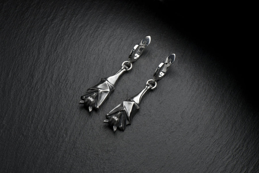 May include: A pair of silver-colored bat-shaped earrings. Each earring features a detailed bat design with outstretched wings and a small loop for attachment. The earrings are suspended from a curved hook. The background is a dark, textured surface. A detailed close-up studio shot of a pair of sterling silver dangle earrings shaped like flying foxes. Each earring features a highly textured sleeping fruit bat hanging upside down, wrapped securely (etsy image id: 7368202736)