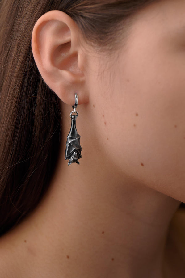 May include: A pair of silver-colored bat-shaped earrings. Each earring features a detailed bat design with outstretched wings and a small loop for attachment. The earrings are suspended from a curved hook. The background is a dark, textured surface. A detailed close-up studio shot of a pair of sterling silver dangle earrings shaped like flying foxes. Each earring features a highly textured sleeping fruit bat hanging upside down, wrapped securely (etsy image id: 7368202736)