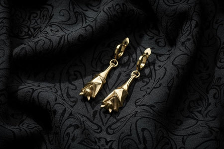 May include: A pair of gold-colored earrings with a unique bat-wing design. The earrings are suspended from a small hoop and feature a detailed, geometric form. The background is a dark fabric with a subtle floral pattern. Reimagined in solid 14k yellow gold, these sleeping flying fox earrings transform from creatures of the night into radiant, sunlit treasures. The warm golden metal highlights the intricate fur and wing details, giving the fruit (etsy image id: 7416093209)