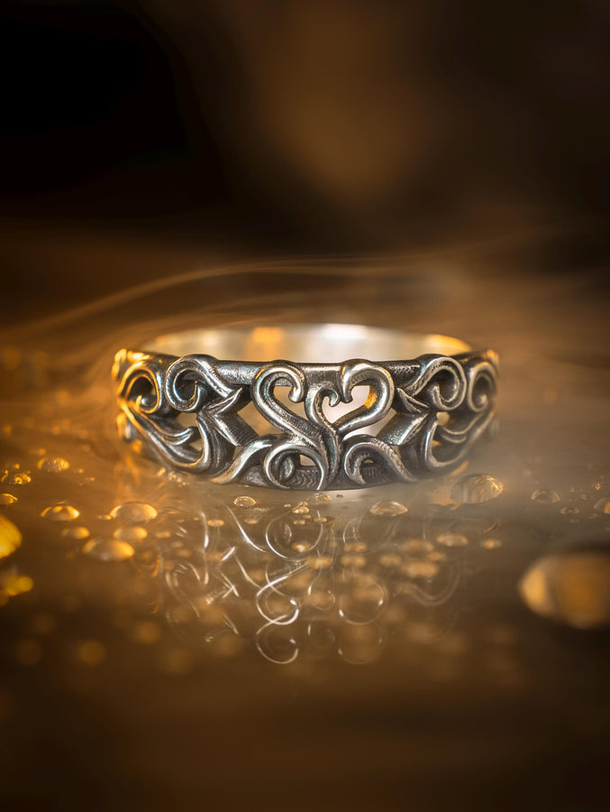 A detailed close-up of a wide sterling silver ring resting on a reflective surface. The band is covered in a deep-relief filigree pattern consisting of interlocking scrolls, vines, and subtle heart shapes. The raised designs are polished bright silver, while the recessed background is heavily oxidized to black, highlighting the complexity of the Baroque motif.