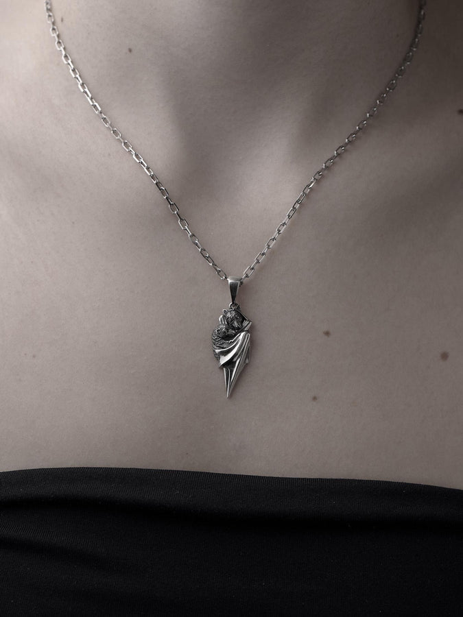 A detailed close-up studio shot of a sterling silver pendant shaped like two flying foxes or fruit bats. The bats are intimately huddled together, wrapped within the smooth, sweeping folds of their leathery wings. The metal is highly textured to show the fine details of their fur and faces. The silver is heavily oxidized in the recesses, creating a striking contrast against the polished highlights of the wings. (etsy image id: 5592925539)