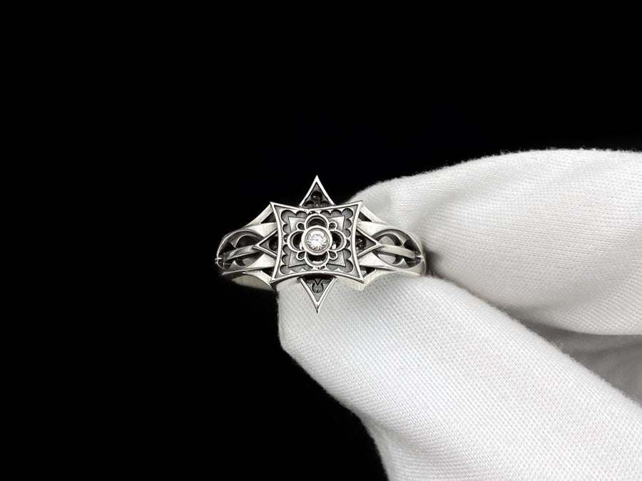 A detailed studio shot of a sterling silver ring held in a gloved hand. The focal point is a geometric "North Star" face—a square frame turned on its point, embellished with top and bottom spikes. Inside the frame is a floral quatrefoil pattern surrounding a round, bezel-set white gemstone (Diamond or CZ). The silver is polished on the raised lines and dark in the recesses. (etsy image id: 7452221522)