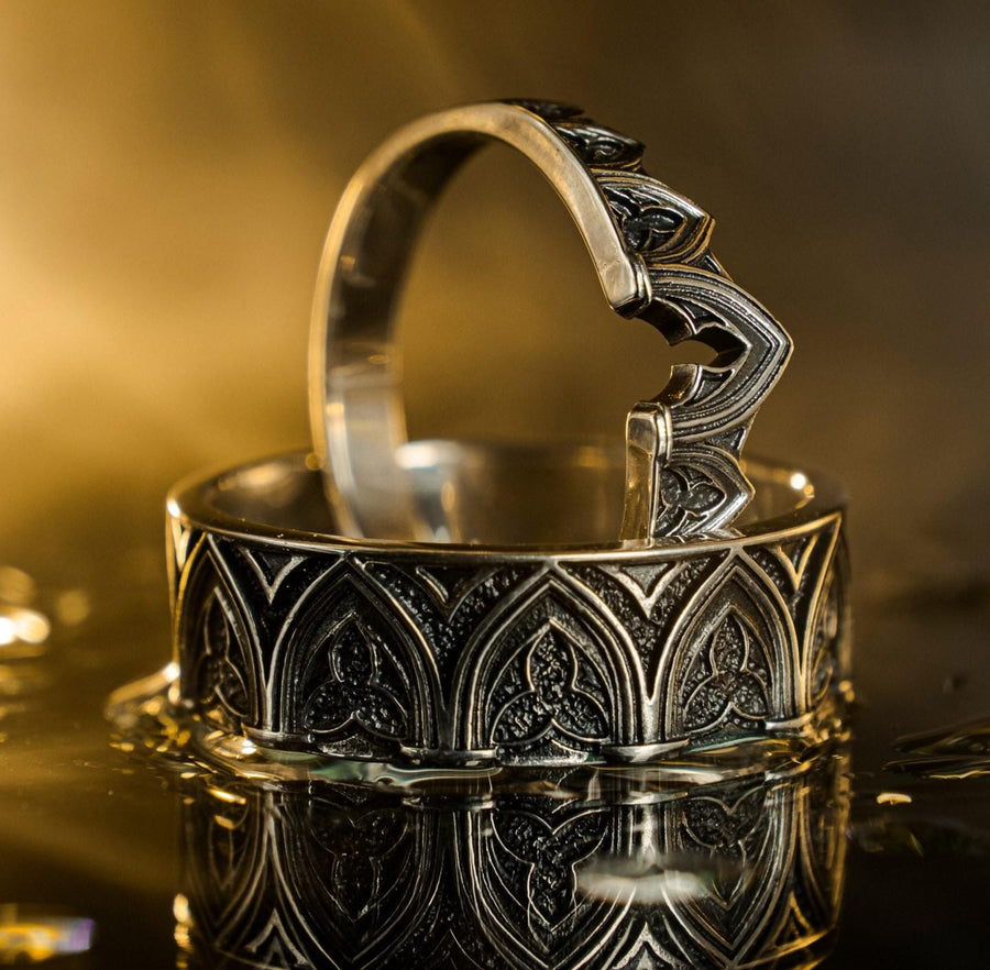 A cinematic studio shot of two matching sterling silver rings. The top ring is a delicate "crown" or tiara shape, formed by the peak of a Gothic arch with trefoil tracery. The bottom ring is a wide band featuring a continuous row of identical arches. The background metal inside the arches has a rough, "eroded" or stippled texture, heavily oxidized to black, while the raised architectural lines are polished smooth.
