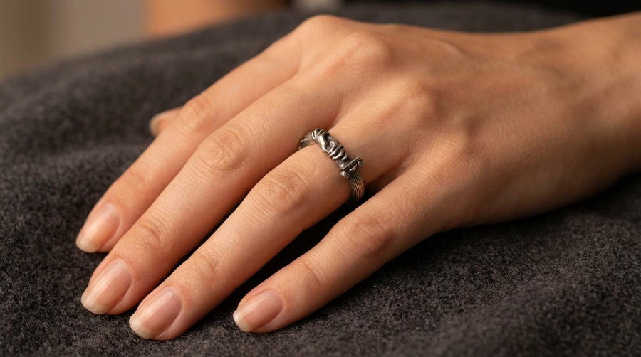 A cinematic studio shot of a sculptural sterling silver ring resting on a dark textured fabric. The focal point is two realistic hands clasped in a handshake (the Fede motif). Instead of fabric sleeves, the wrists emerge from architectural column capitals with distinct "volute" scrolls (Ionic style). The band continues as a textured, fluted column shaft. The silver is heavily oxidized to mimic ancient stone. (etsy image id: 7452415972)