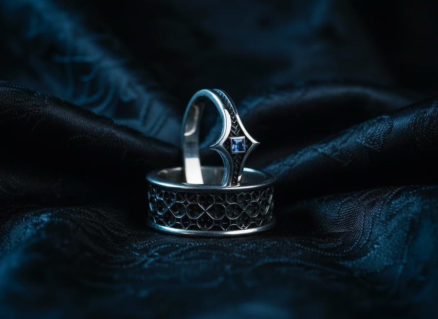 A high-definition shot of a matching two-piece wedding ring set resting on dark velvet. The foreground features a delicate sterling silver ring with a pointed, kite-shaped face holding a square blue gemstone. Standing behind it is a wide, sturdy band with a repeating quatrefoil "clover" pattern. Both rings share a cohesive finish: bright polished silver highlights against a deeply oxidized, matte black background.