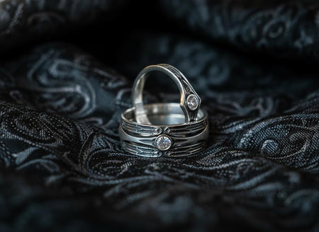 A high-contrast studio shot of a two-piece sterling silver ring set. The foreground features a wide band lying flat, set with a round clear gemstone. Flanking the stone are horizontal, grooved lines resembling arrows or spears. Standing behind it is a narrower engagement-style ring with a matching "lance-tip" shank leading to a central bezel-set white stone. The background is a textured, dark fabric.
