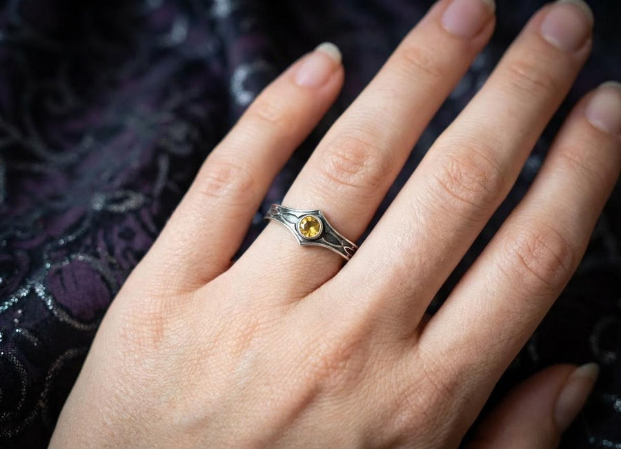 A cinematic close-up of a sterling silver ring with a distinct "V" shaped or "Lance Tip" contour. The focal point is a bezel-set, round gemstone with a deep amber-orange hue (Citrine or Madeira Citrine). The shoulders of the band feature horizontal grooves that taper toward the center, resembling the shaft of a spear or the fletching of an arrow. The metal is polished but features dark oxidation in the grooves for contrast. (etsy image id: 7489049399)