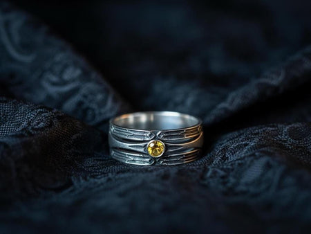 A detailed studio shot of a wide, heavy-gauge sterling silver ring. The focal point is a round, deep red gemstone (Garnet) bezel-set in the center. Flanking the stone are three parallel, tapered grooves that resemble the fletching of an arrow or the futuristic paneling of armor. The metal surfaces are brushed silver, while the deep grooves are blackened with oxidation to emphasize the directional lines. (etsy image id: 7441158070)