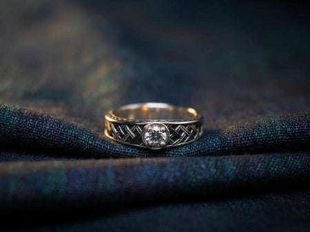 A close-up studio shot of a sterling silver ring featuring a traditional Celtic "basket weave" or knot pattern. The strands of silver appear to weave over and under each other in a continuous loop. The center of the ring holds a round, clear white gemstone (Diamond/CZ/Moissanite) in a secure bezel setting. The recesses of the weave are darkened with oxidation to create high contrast against the polished silver strands. (etsy image id: 7441489606)