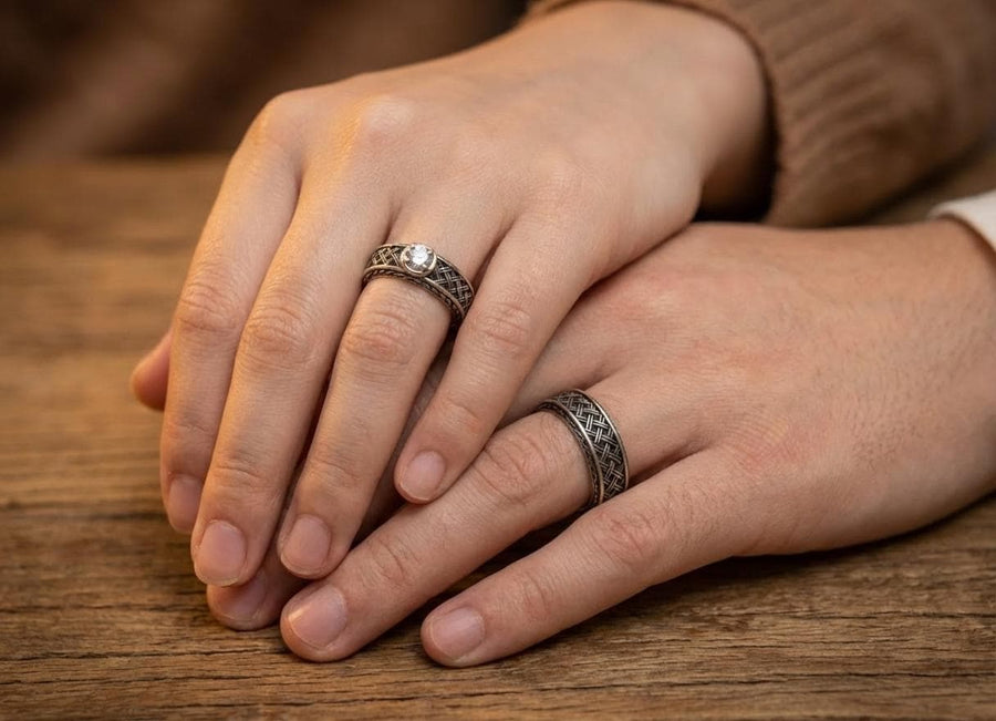 A warm, atmospheric shot of a matching wedding ring set. The engagement ring stands upright, showcasing a round white gemstone in a bezel setting. Lying flat beneath it is a matching wide band without a stone. Both rings share a cohesive design: a central diagonal "basket weave" pattern flanked by twisted rope borders. The silver is oxidized to reveal the intricate texture of the weave.