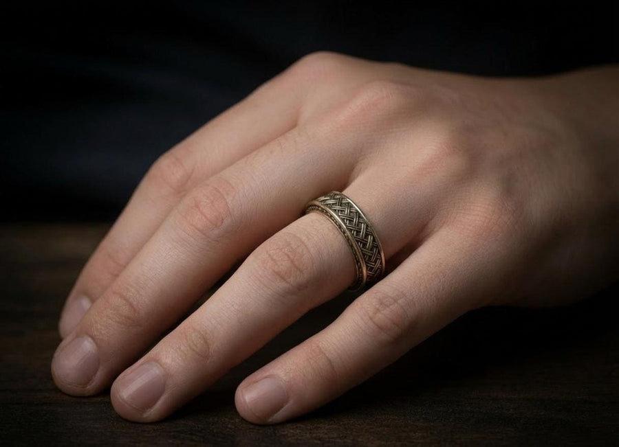 A cinematic studio shot of a wide sterling silver ring lying on a reflective golden surface. The band features a central "basket weave" pattern—diagonal strands of silver crossing over and under each other. The edges are framed by a distinct "rope" or "cable" twist border. The raised metal is polished silver, while the deep recesses are blackened to highlight the intricate weaving.