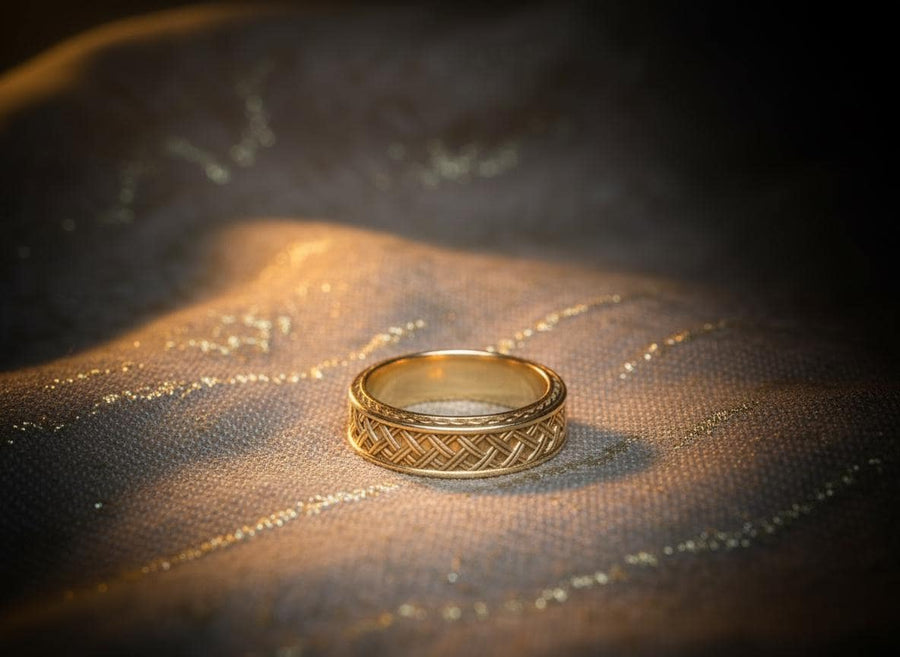 A cinematic studio shot of a wide sterling silver ring lying on a reflective golden surface. The band features a central "basket weave" pattern—diagonal strands of silver crossing over and under each other. The edges are framed by a distinct "rope" or "cable" twist border. The raised metal is polished silver, while the deep recesses are blackened to highlight the intricate weaving.