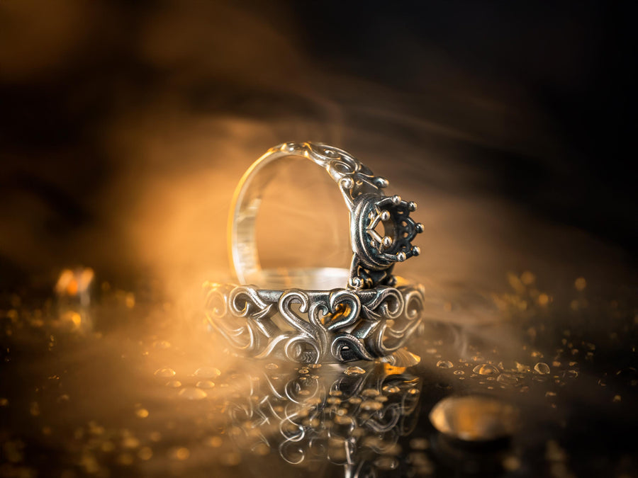 A high-contrast studio shot of a two-piece sterling silver ring set. The top ring features a raised, three-dimensional "crown" or "coronet" motif with small spheres on the tips. The bottom ring is a matching wide band. Both rings are covered in intricate, raised scrollwork or vine-like filigree. The silver is polished bright on the raised swirls, contrasting with the black oxidized background.