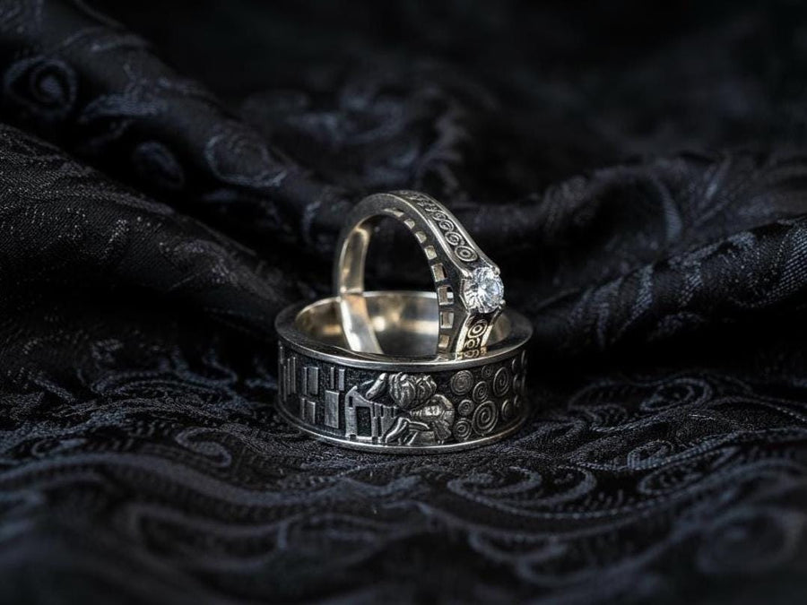 A cinematic studio shot of a matching two-piece ring set. The bottom ring is a wide silver band featuring a detailed relief carving of the couple from Gustav Klimt's "The Kiss." The top ring is a narrower engagement band that mimics the geometric patterns of the painting and holds a round, amber-colored gemstone. The background is a warm, golden haze, reflecting the "Golden Phase" of the original artwork.