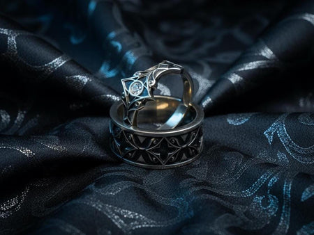 A cinematic studio shot of a matching two-piece ring set. The top ring is a delicate "Tiara" style band featuring a lozenge-shaped centerpiece with a round red Garnet. The bottom ring is a wide band with a repeating geometric "Star" or "Cross" tracery pattern. Both rings share a cohesive oxidized sterling silver finish, with polished highlights accentuating the intricate relief work against a dark background.