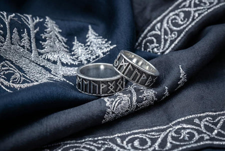 A clean studio shot of two matching sterling silver rings. The bands feature the 24 characters of the Elder Futhark runic alphabet wrapping around the exterior. The runes are polished silver, standing out sharply against a rough, blackened background texture. The rims are smooth and polished, framing the ancient script.